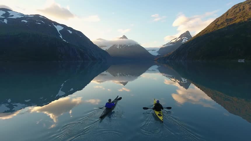 Two kayakers paddling on a still body of water that is reflecting the mountain scene in front of them