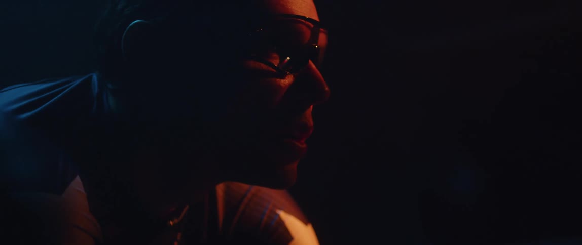 Man wearing glasses and top with white star on shoulder raises his head in dark area