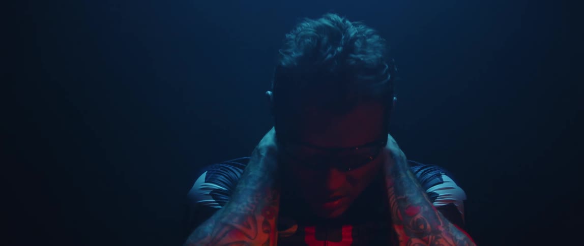 Tattooed man holds his hands on his neck under blue and red lights inside a dimly lit room