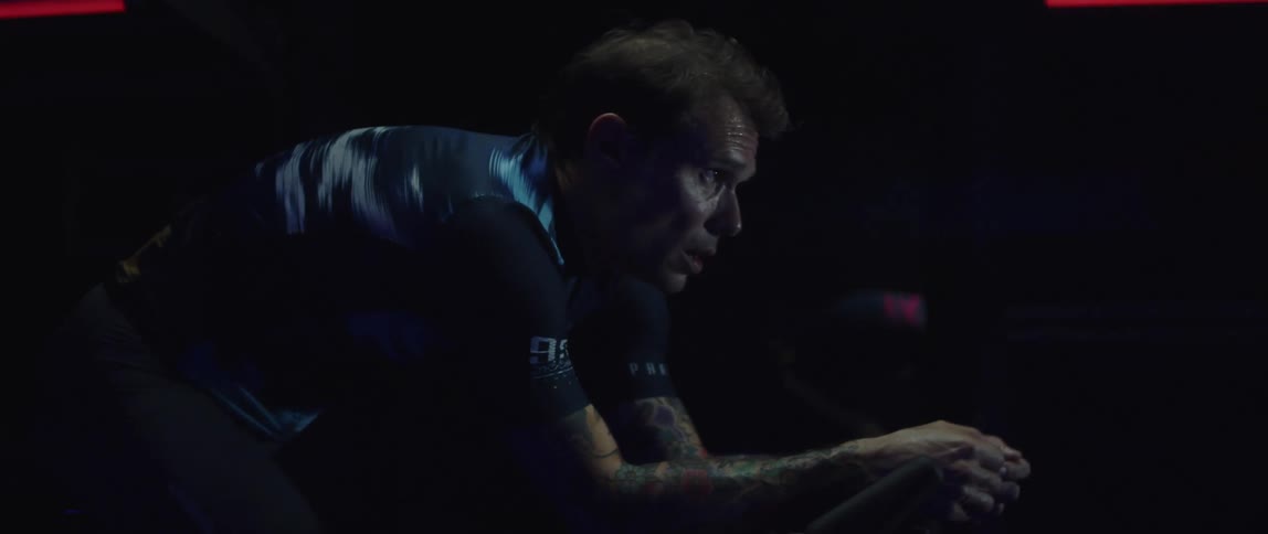 Focused sweaty man exercising on a stationary bike next to a mirror in a dark gym room