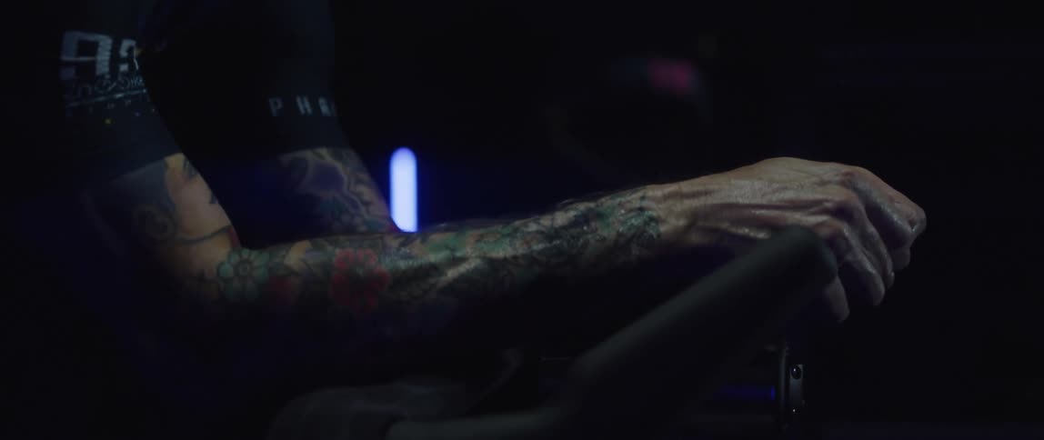 Tattooed man pedaling on a static bicycle inside a dimly lit room