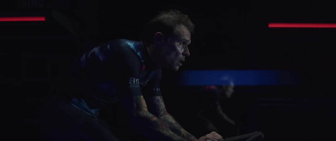 A man with tattoos rides an indoor bicycle in a dark room
