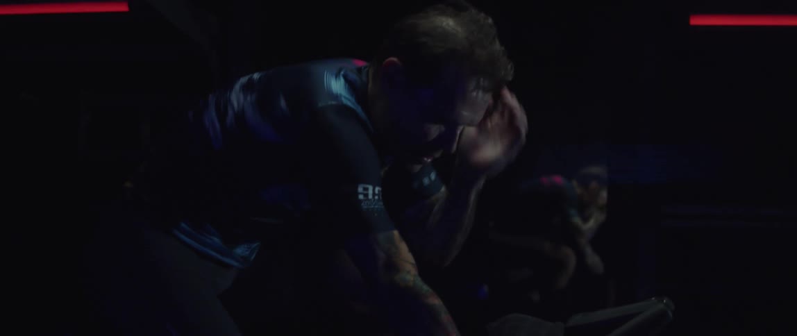 A man wipes his neck and chin with his hand while exercising on a exercise bike in a dark room