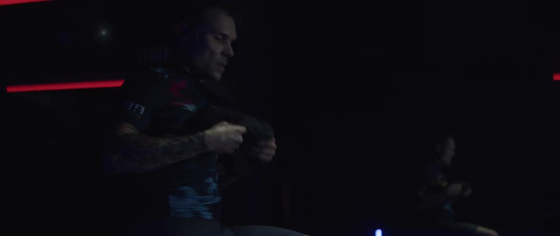 A man exercising on a stationary bike picks up a towel and wipes the sweat off his face and then runs his fingers through his hair
