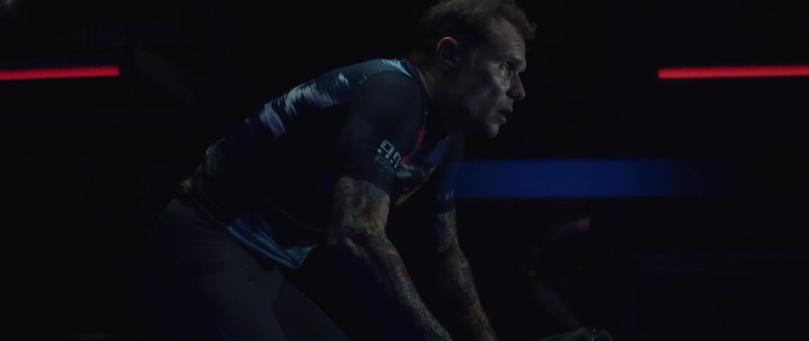 A tattooed man riding a stationary bike in a dark room