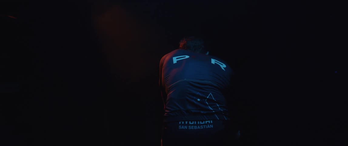 Back of a man riding a static bicycle inside a dimly lit room