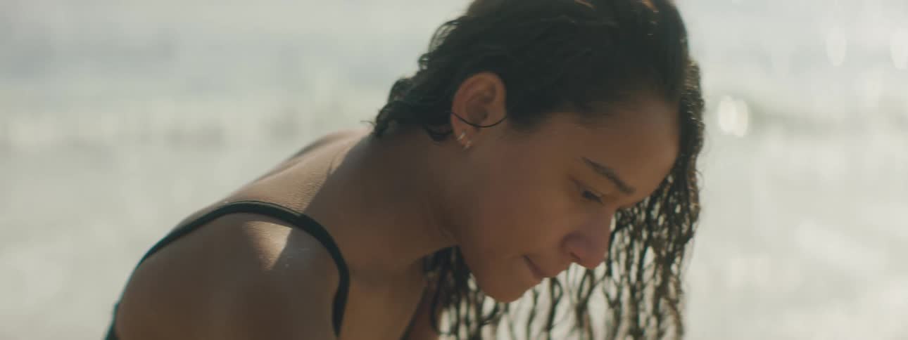 A young woman sitting in the sands of a beach on a sunny day turns her head to look behind her