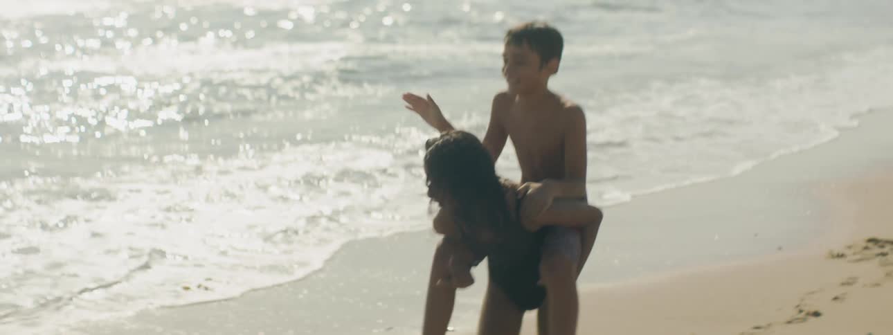 A girl wearing a swimsuit carries her brother on her back along the beach during the day