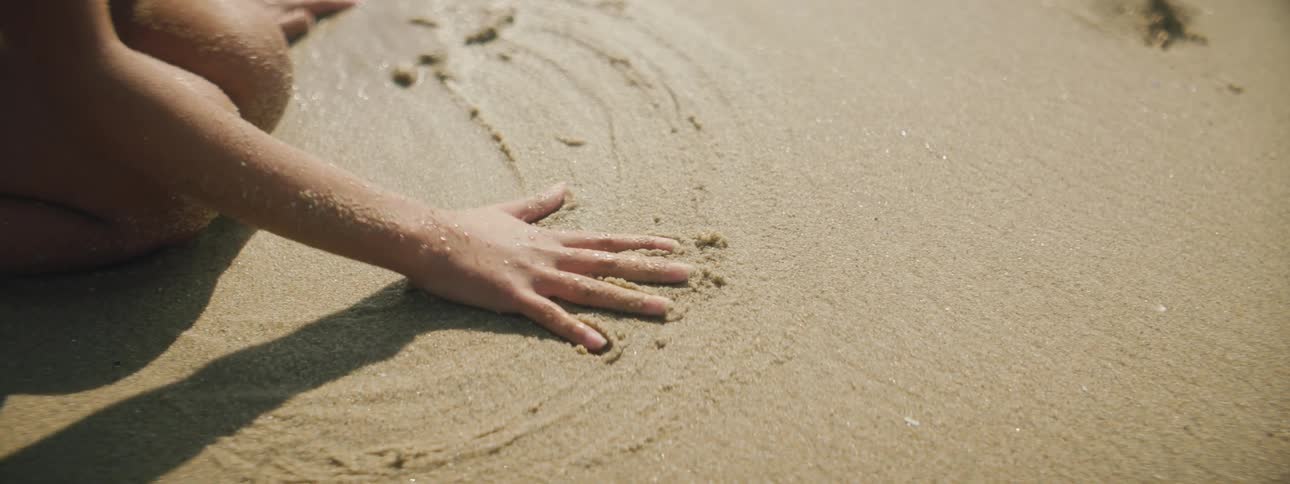 A girl makes a handprint in sand while kneeling on a wet beach