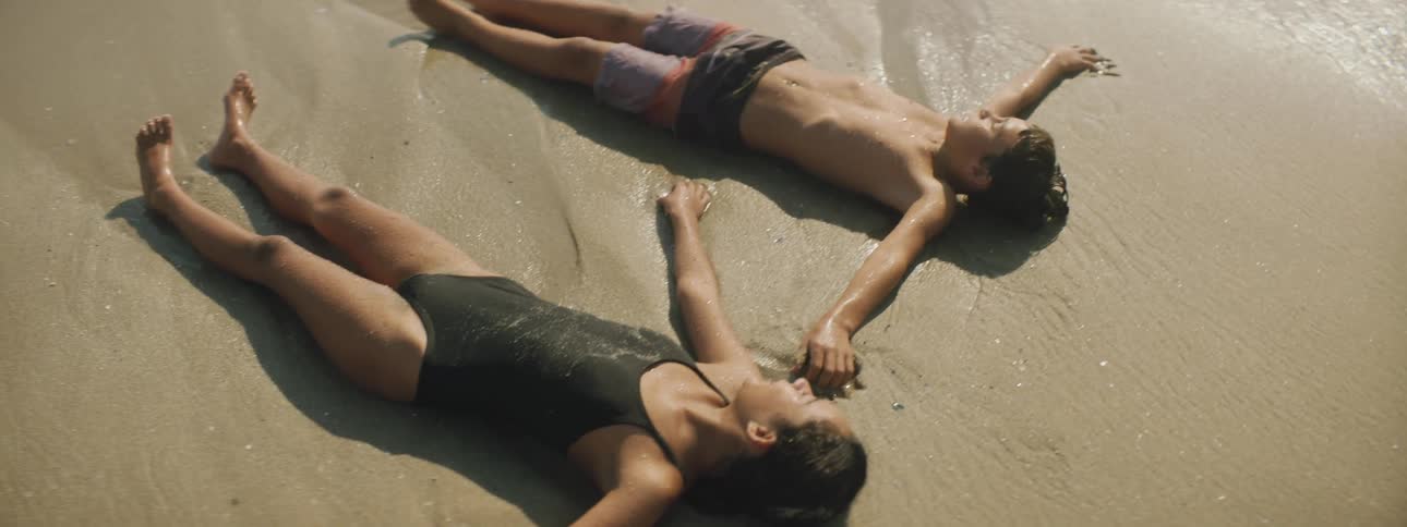 A young boy and girl lay on the sandy beach with the ocean water coming into to them