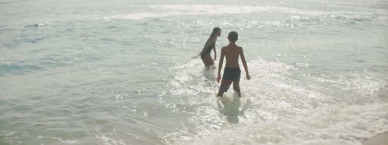 A brother and sister push and splash one another as they play in the water on the shore