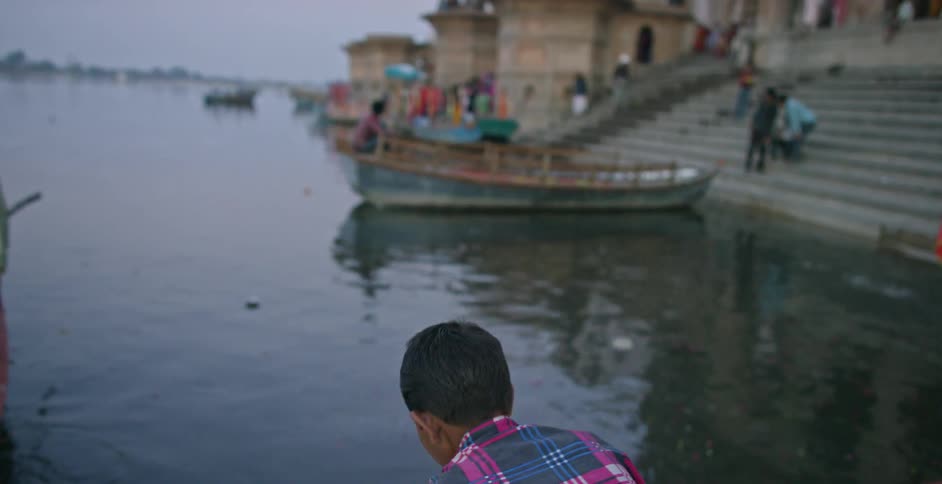 A boy approaches a river with a lit candle and moves to place it in the water
