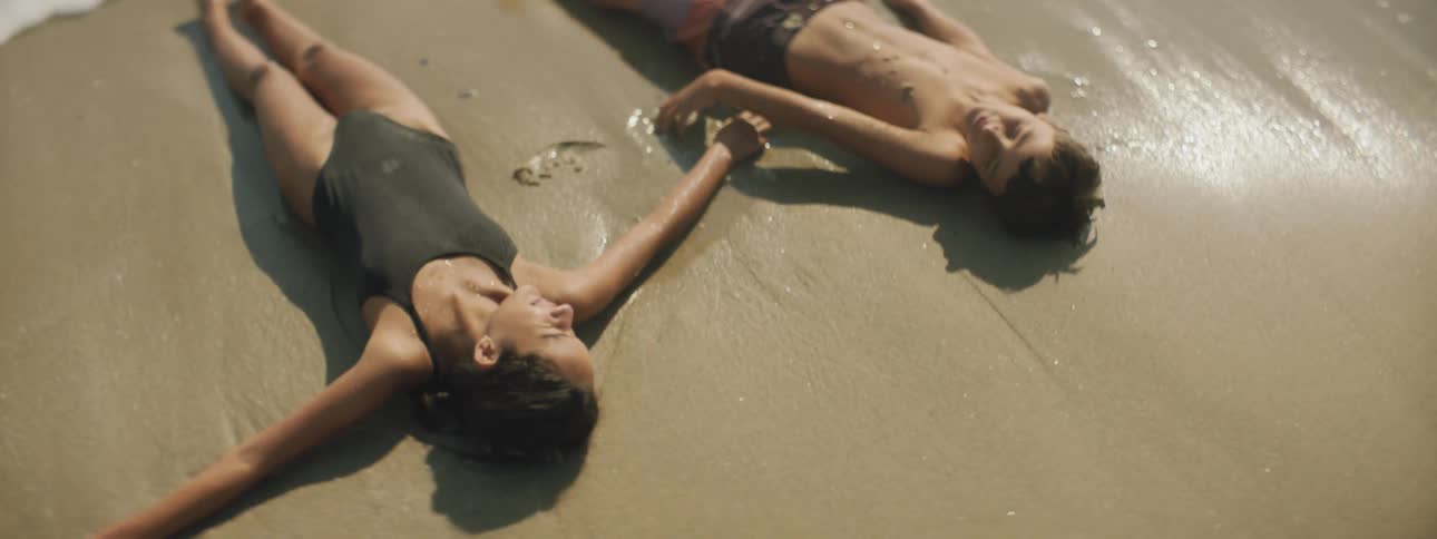 A girl and boy lay on the beach as the ocean waves crash up on to them
