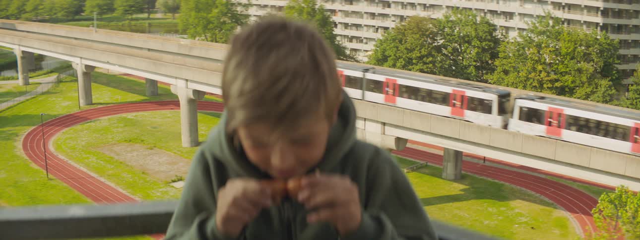 A boy eats a chicken wing on his balcony as a passenger train passes by behind him