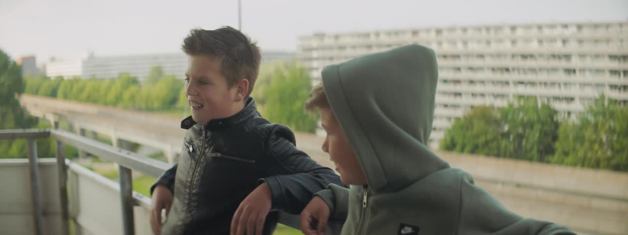 Two young white boys stand on a balcony and talk and laugh with one another