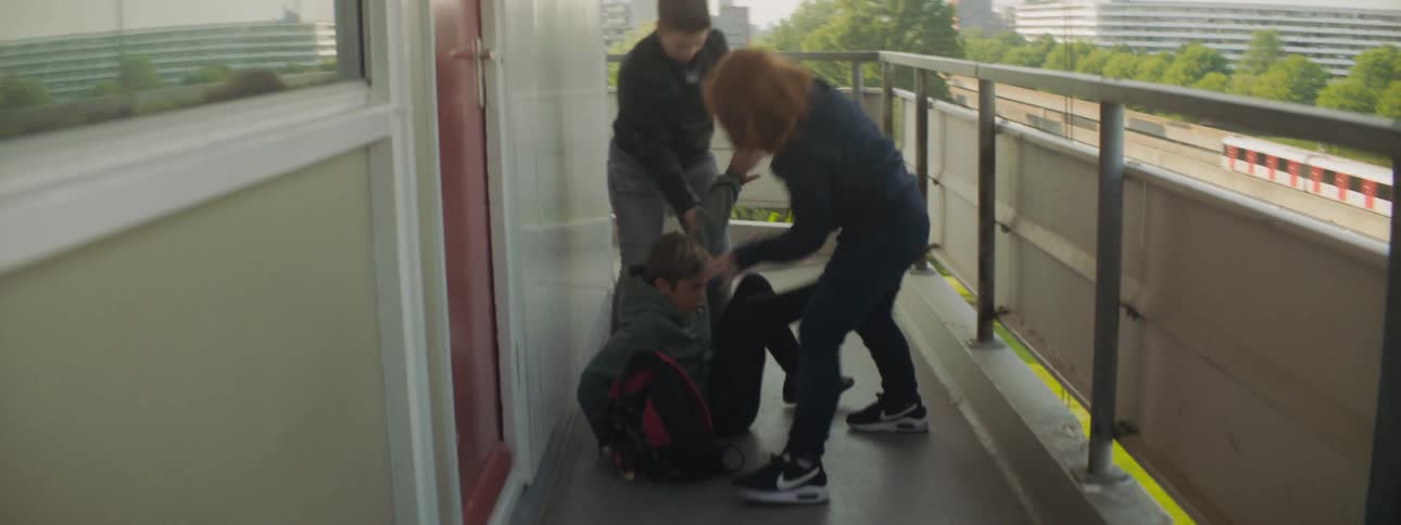 A young man and woman kick and take the belongings of a boy sitting on a balcony in a city during the day