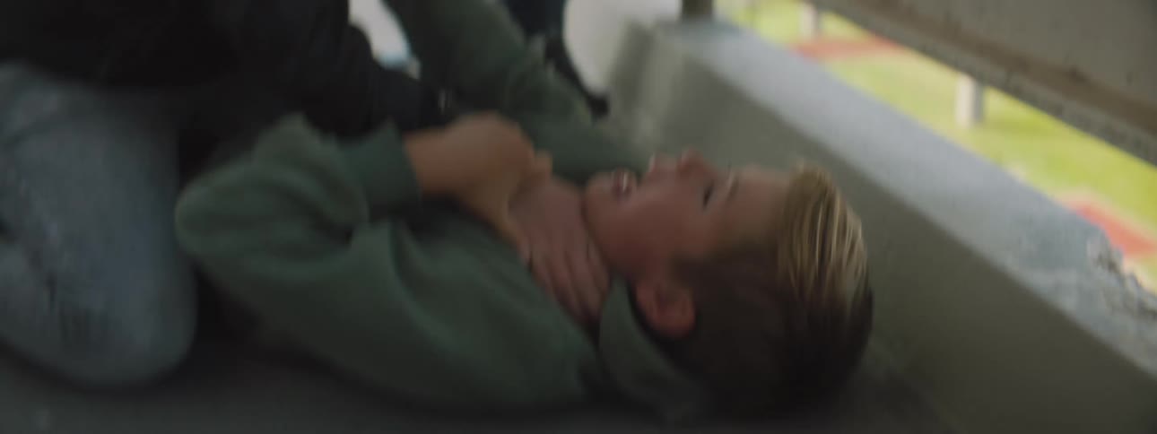 A young boy knees on another boy and puts his hand around his neck to choke him as another boy stands by