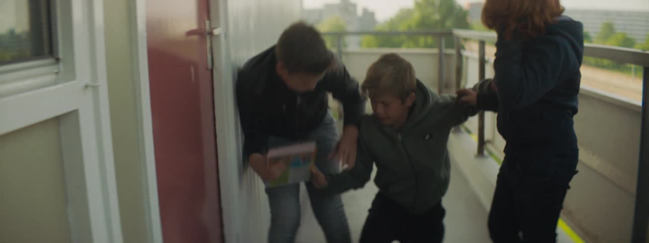 A boy climbs to his feet and attempts to pursue another boy who is stealing his books but is physically restrained by a third boy