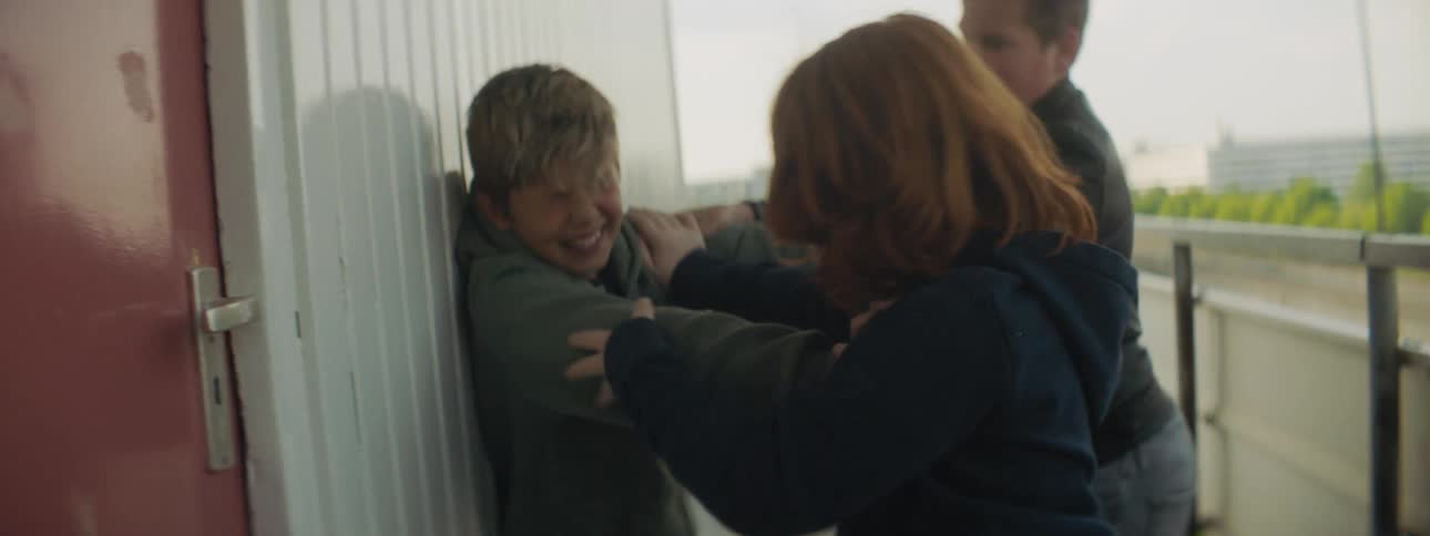 Two boys are holding another boy against a wall next to a railing as he struggles and they hit him