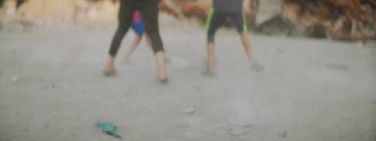A few people play soccer on a dirt ground with rocks in the background