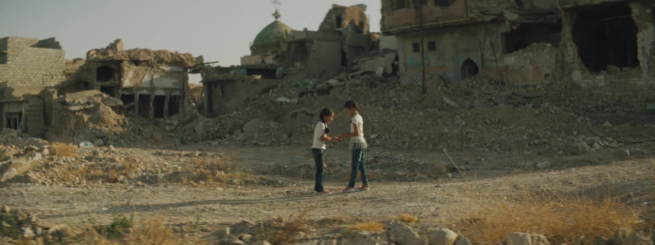 Two young girls play a clapping game as they stand in front of debris and destroyed homes in their village