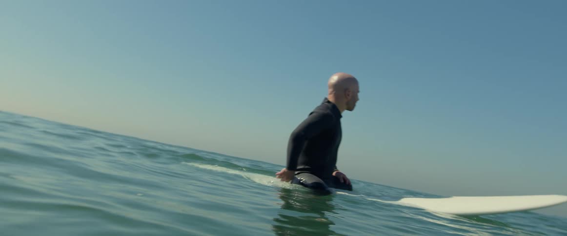 A man wearing a black wetsuit is in the water sitting on his surfboard