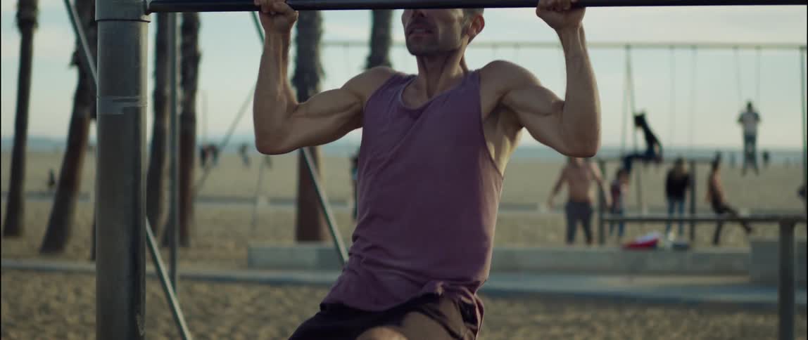 A man does pull ups at a park on the beach while others swing and walk behind him