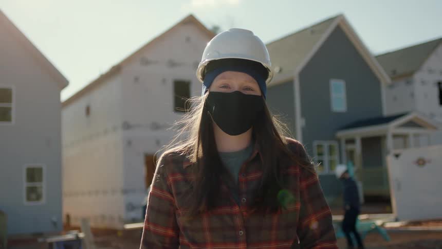A woman wearing a hardhat and cloth mask stands in front of newly constructed and repaired homes