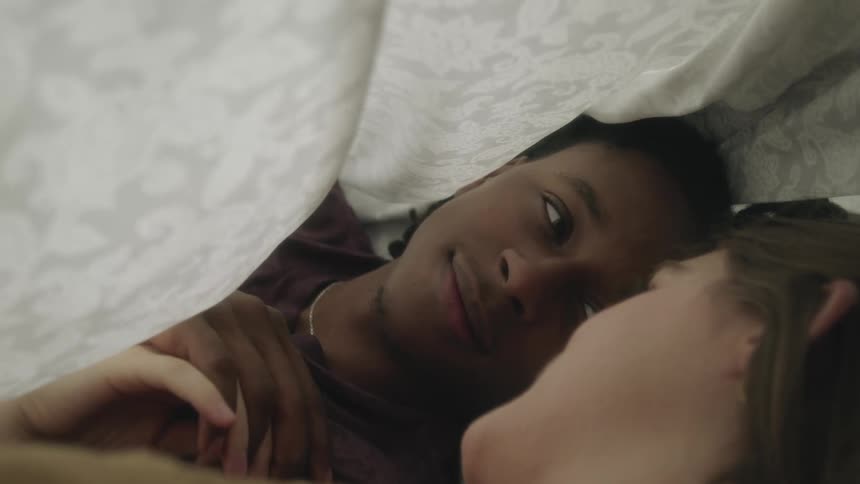A couple hold hands and laugh while lying under a sheet in bed