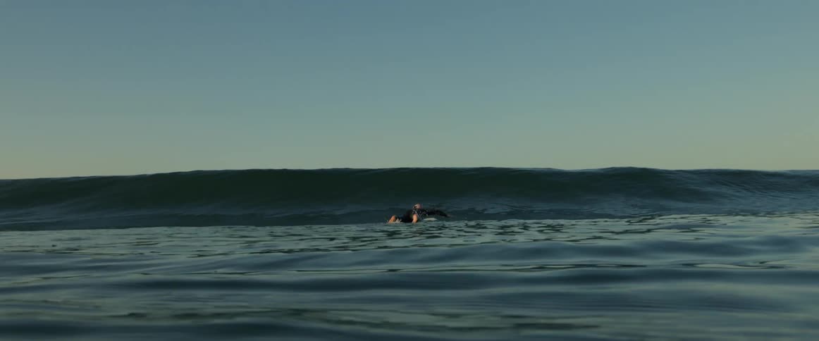 A surfer wearing a black wetsuit lies on his surfboard and paddles with his hands as he goes over a wave