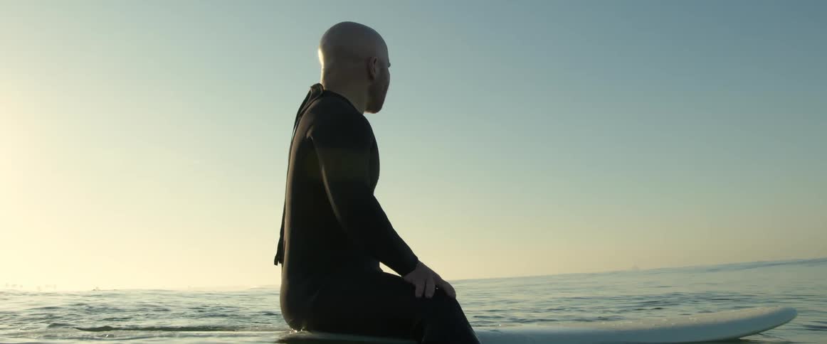 A man wearing a black wetsuit is in the water sitting on his surfboard and looking around