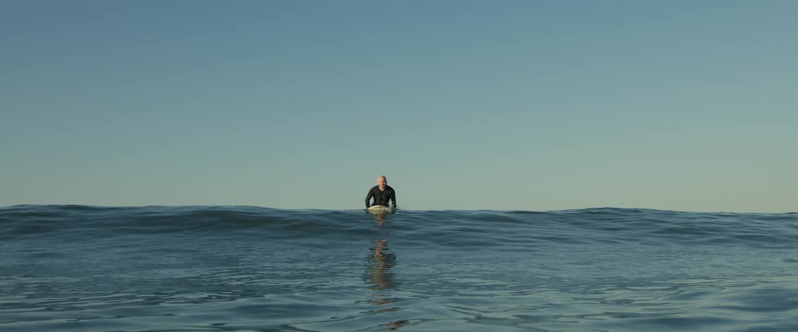 A surfer wearing a black wetsuit lies on his surfboard and raises himself up as a wave grows