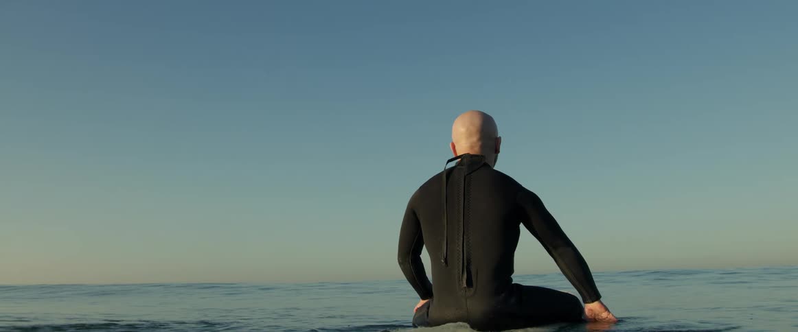 An older white man is sitting on his surfboard on the ocean