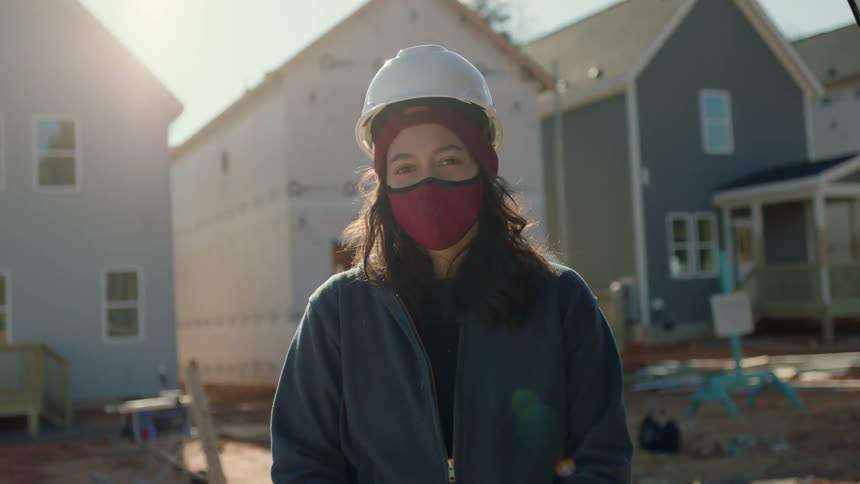 Woman wearing a hard hat and face mask stands in front of a row of houses under construction