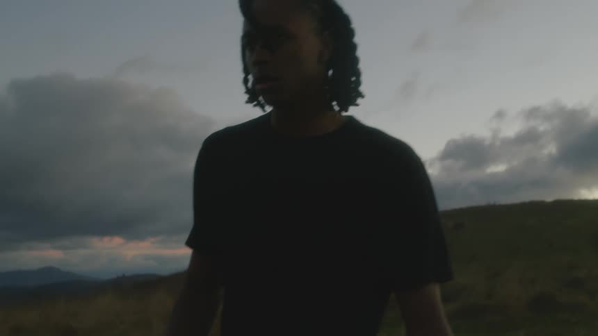 A black man is walking outside under a cloudy sky and there are mountains in the distance