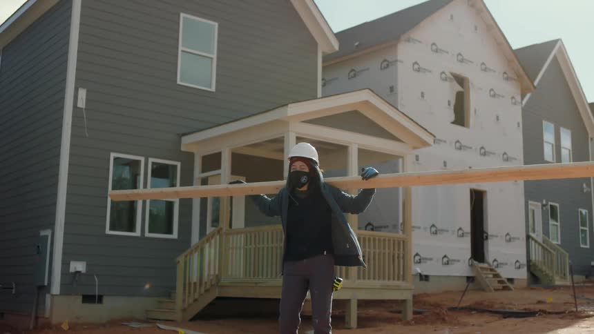 Female construction worker wearing a white hardhat carries a large wood plank over her shoulders