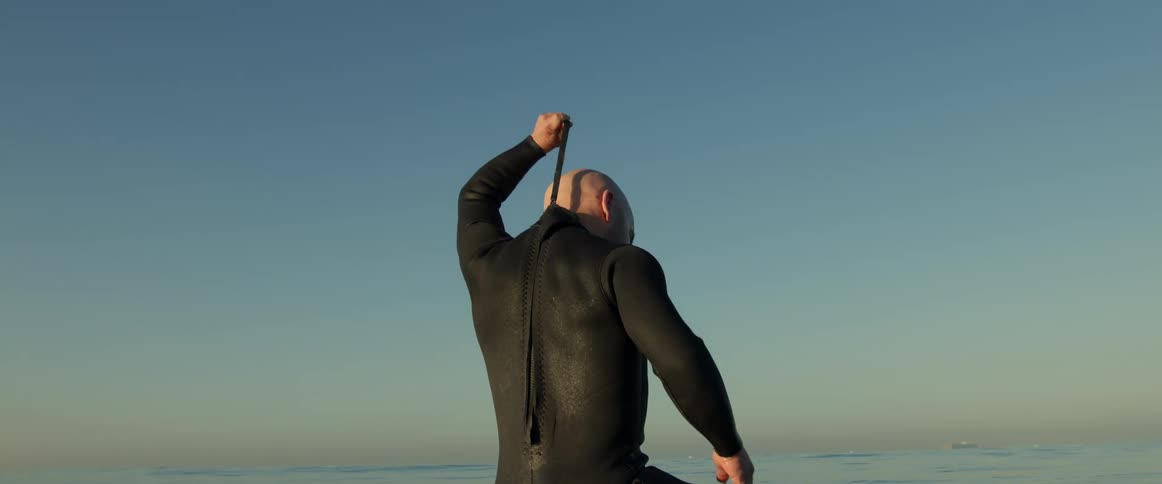 A man pulls on a strap on his wet suit as he sits on a surf board in the ocean