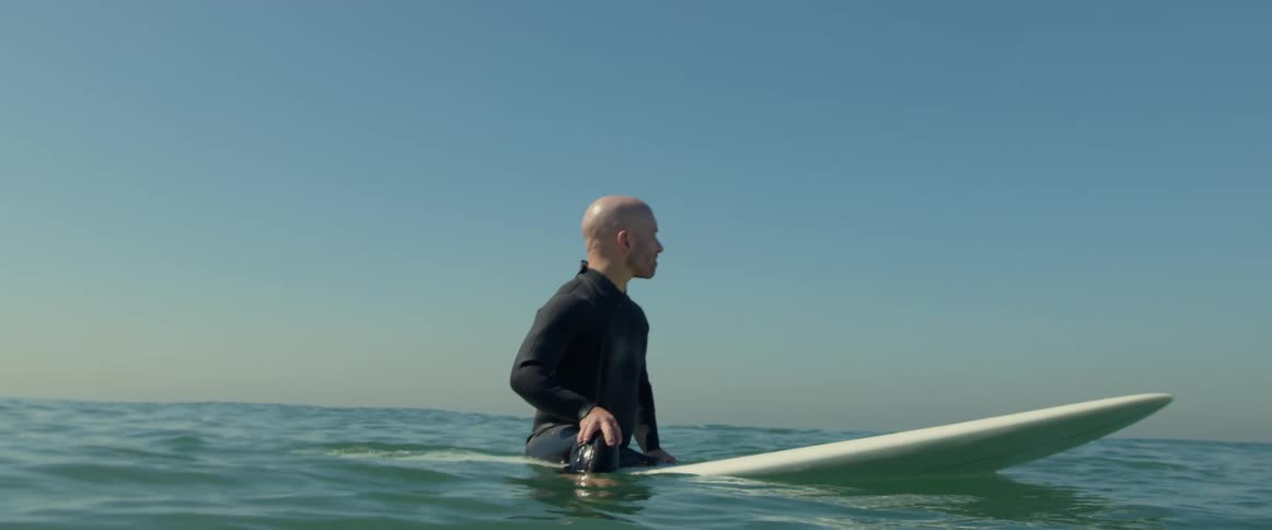 A man is sitting on his surfboard with his hand at his waist as he waits for a wave