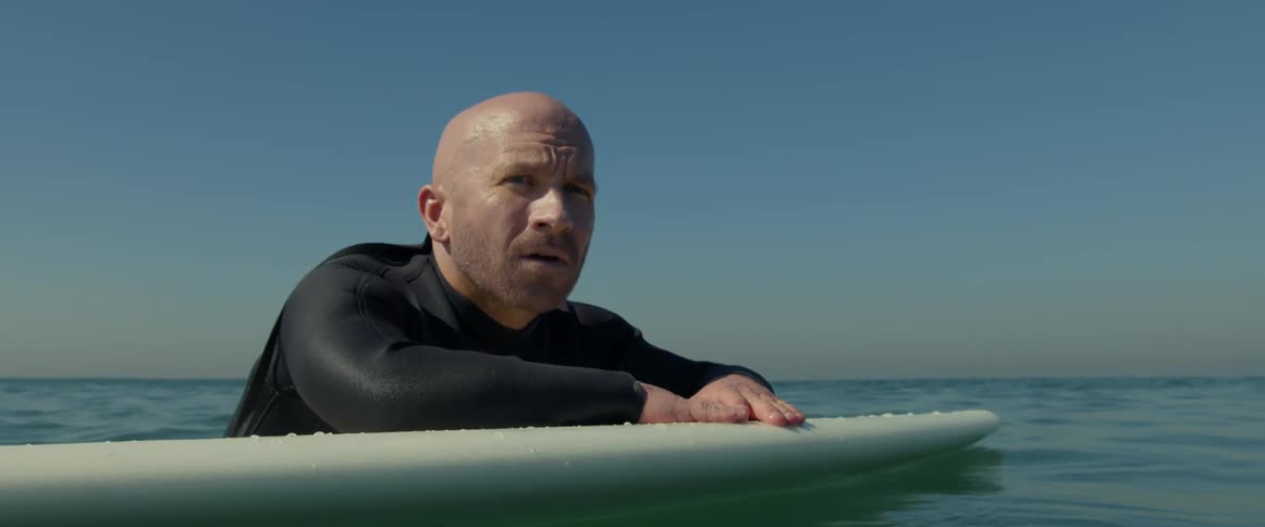 A surfer wearing a black wetsuit is floating on his surfboard and looking around