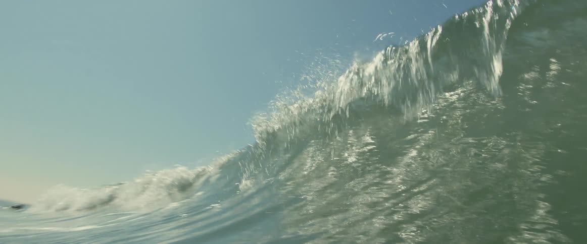 Large green tinted wave crests and crashes under blue sky