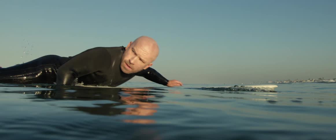 A surfer wearing a black wetsuit lies on his surfboard and paddles with his hands