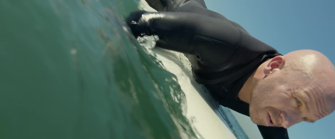 A surfer wearing a black wetsuit lies on his surfboard and paddles with his hands