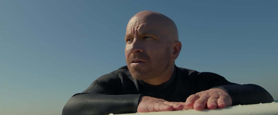 A surfer wearing a black wetsuit is floating on his surfboard and looking around