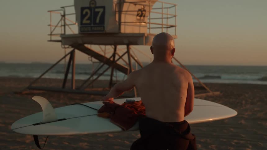 An older white man is walking on the beach and holding his surfboard