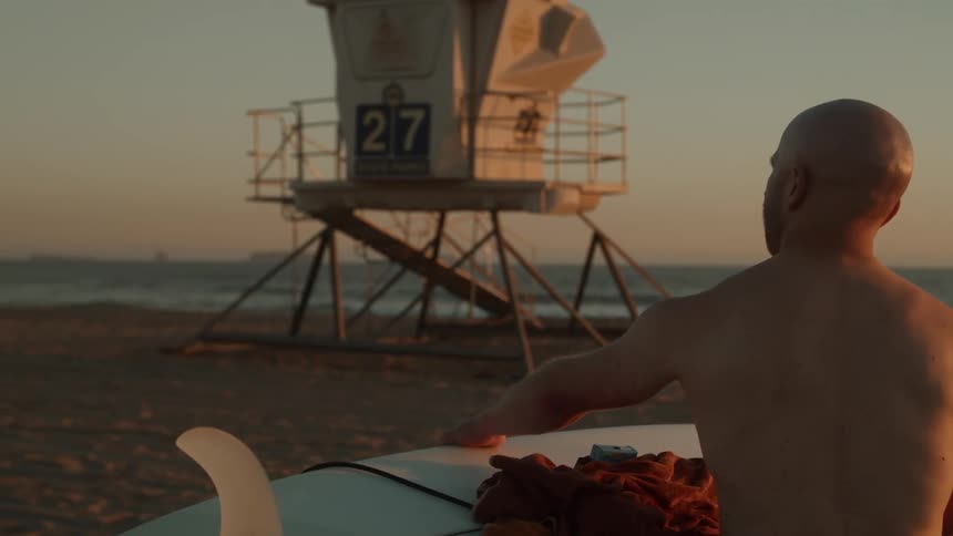 A man carrying his surfboard toward the ocean waves and lifeguard tower