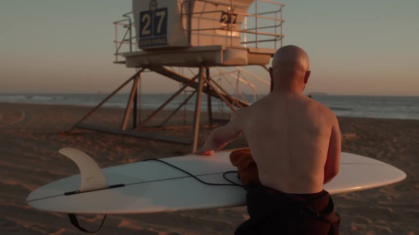 A man is walking across a beach past a lifeguard stand holding his surfboard
