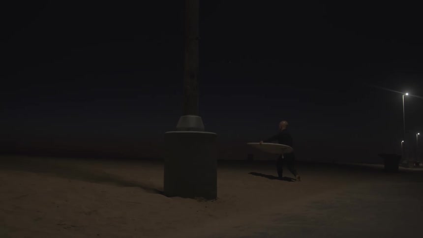Man wearing wet suit carries surfboard up sandy slope at night