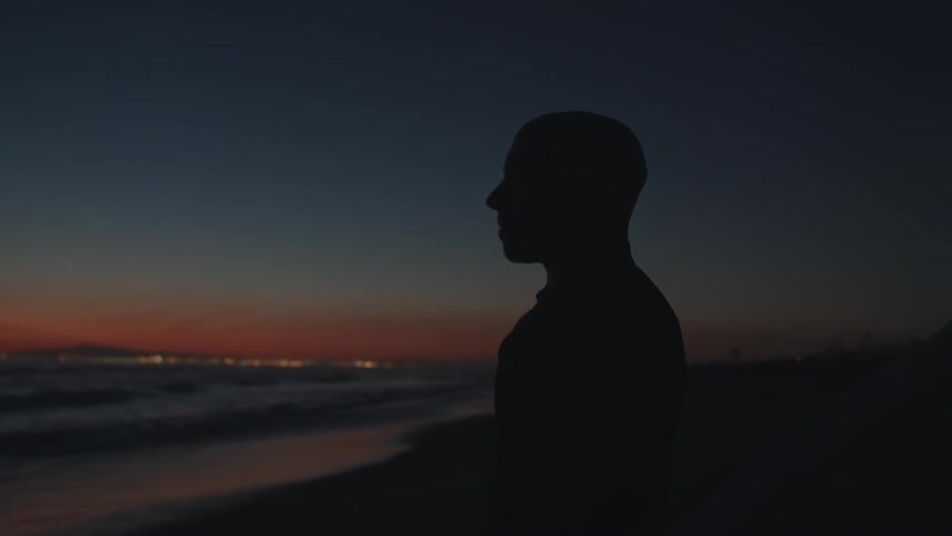 The silhouette of a man with his surfboard looking out at the ocean as night falls