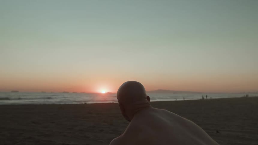 An older white man is holding a surfboard on the beach at sunset