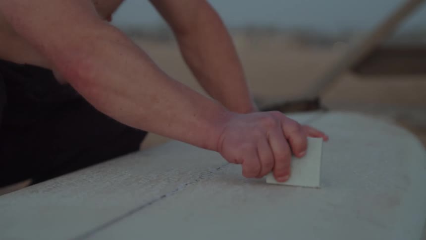 A man sits on the sand and rubs his surf board with wax